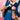 A person in a blue Venchi uniform holds an ice cream cone with a scoop of pink ice cream and uses a metal ice cream scooper, against a pink background. - AI generated text