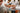 People in white lab coats examine and reach for a white tray filled with neatly arranged pieces of chocolate in a bright, indoor setting. - AI generated text